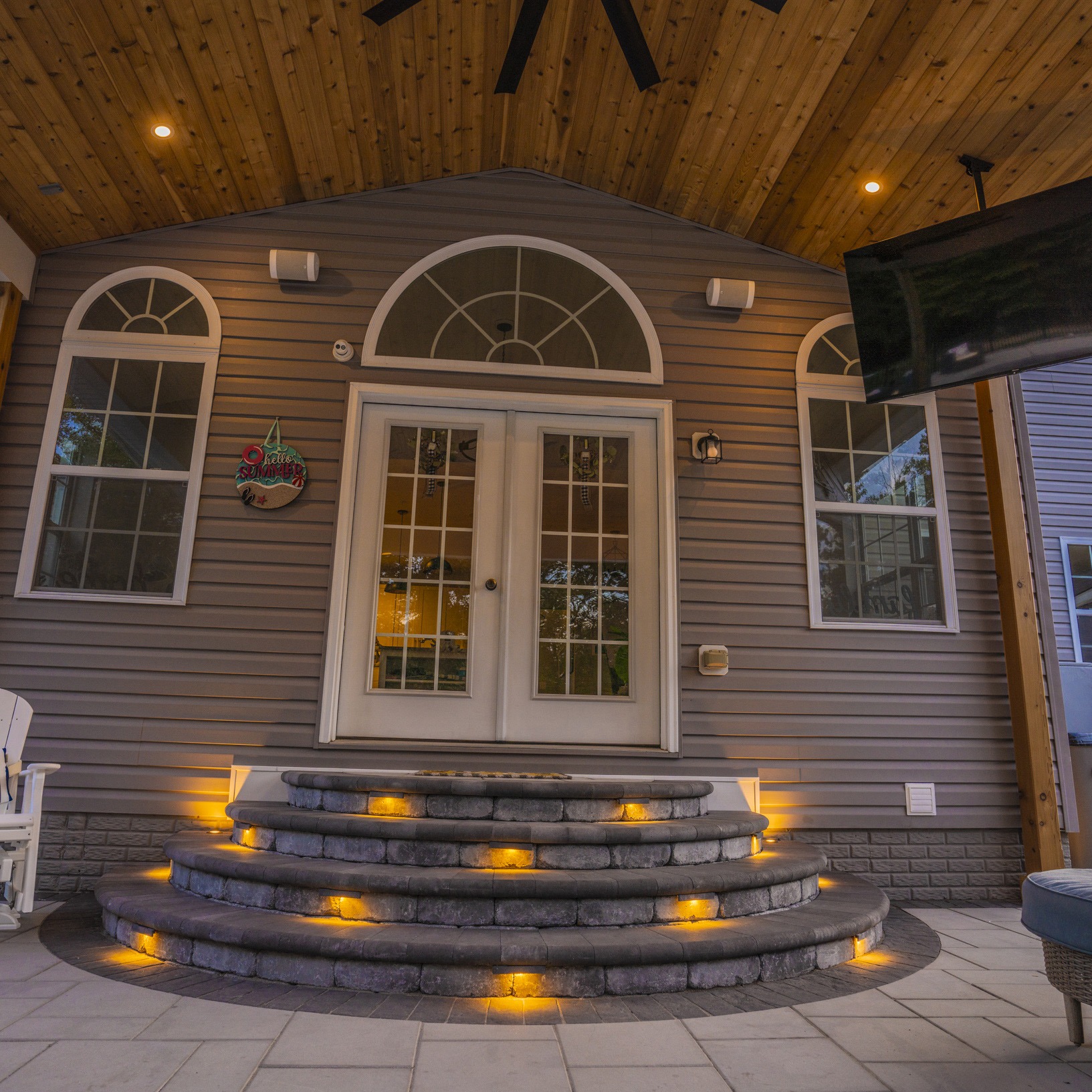Cozy outdoor patio with lit steps, arched windows, and double doors. Ceiling fan overhead. Relaxed, inviting atmosphere for summer gatherings.