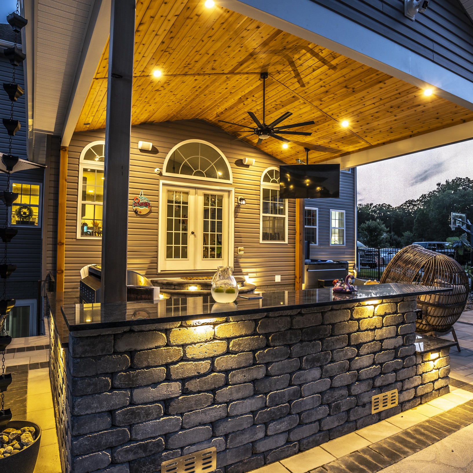 Cozy outdoor patio with stone bar, wooden ceiling, ambient lighting, wicker seating, and glass doors leading into a modern suburban home.