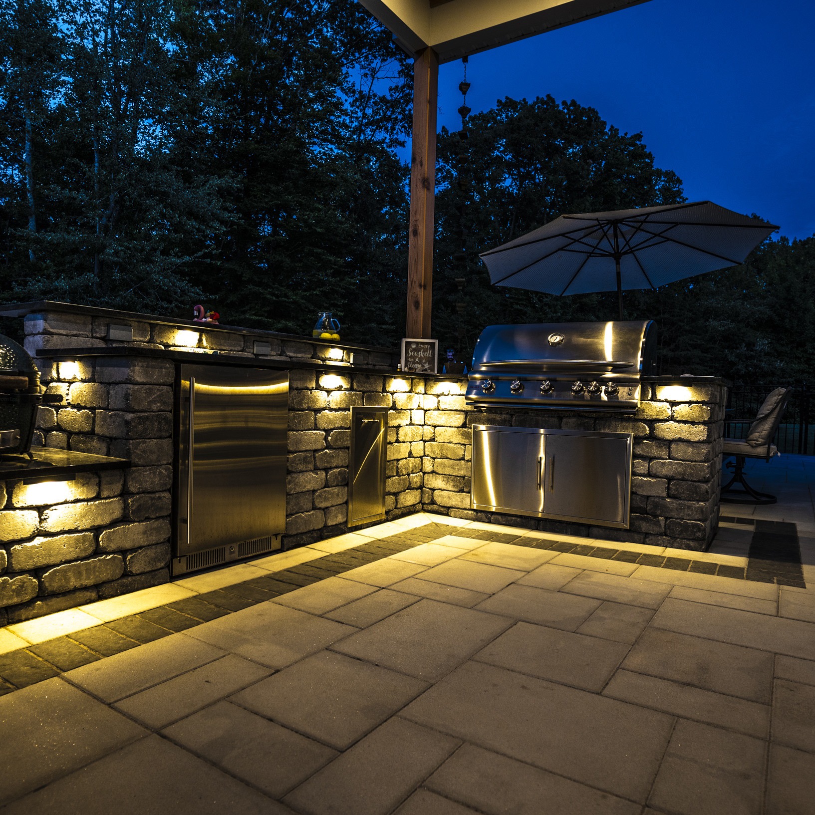 An outdoor kitchen with stainless steel appliances under a wooden shelter, illuminated by soft lighting, surrounded by trees during twilight.