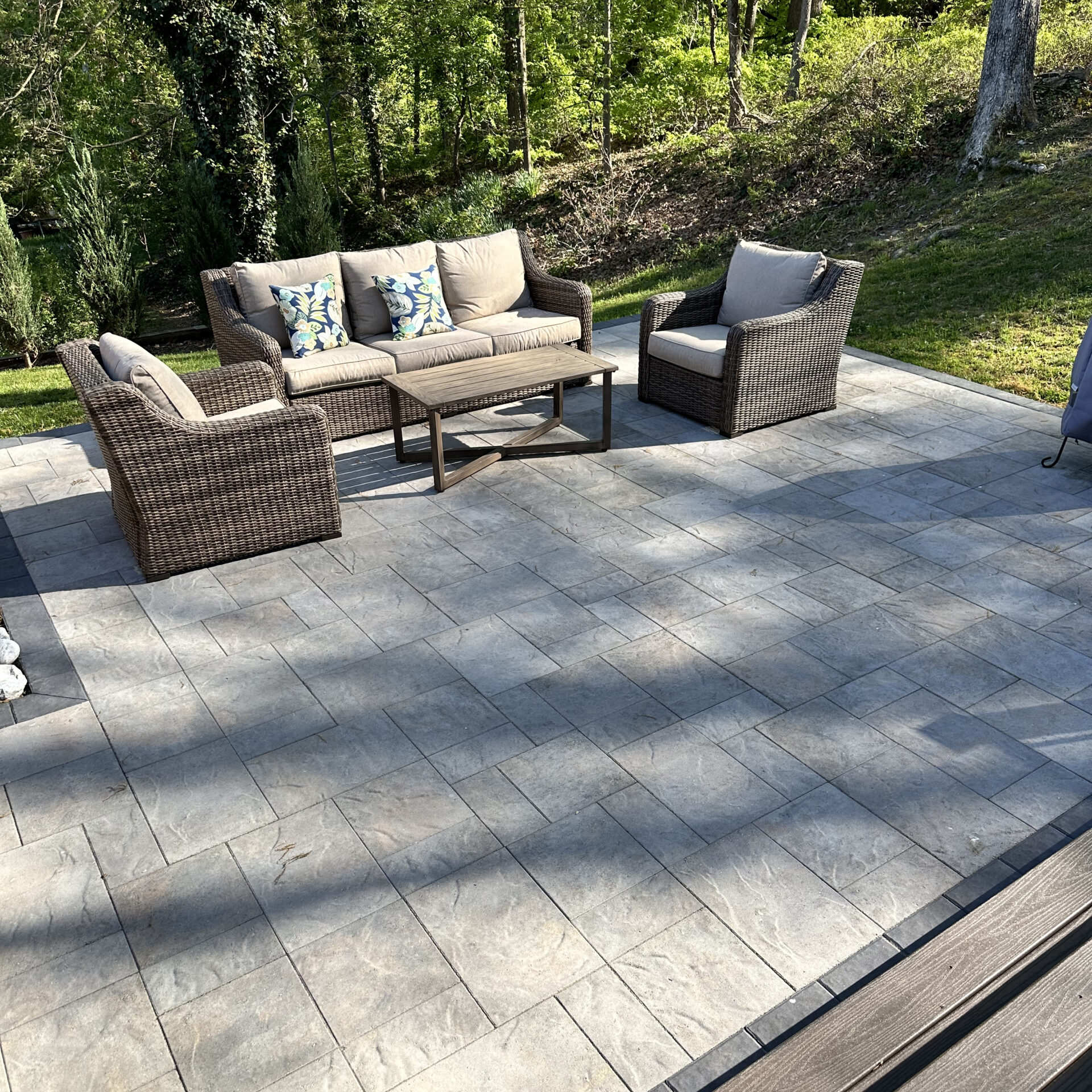 A serene patio with cushioned wicker furniture set on gray stone pavers, surrounded by lush green trees and nature.