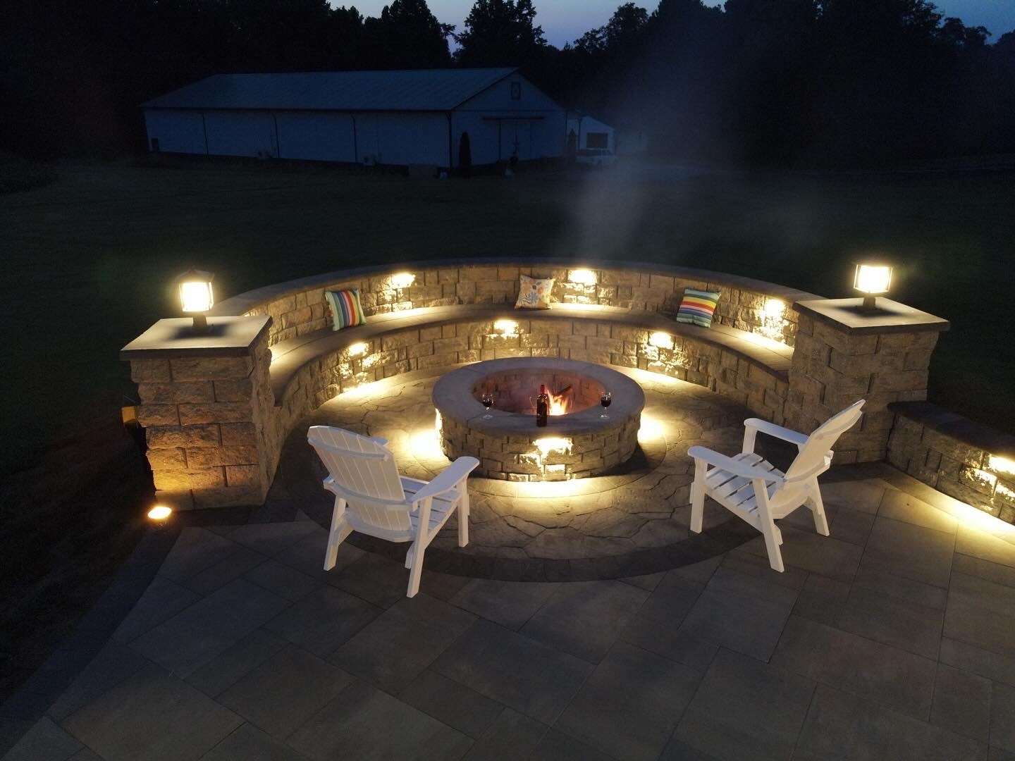Circular stone fire pit area with white chairs and colorful cushions, surrounded by soft lighting. Large building in the background.