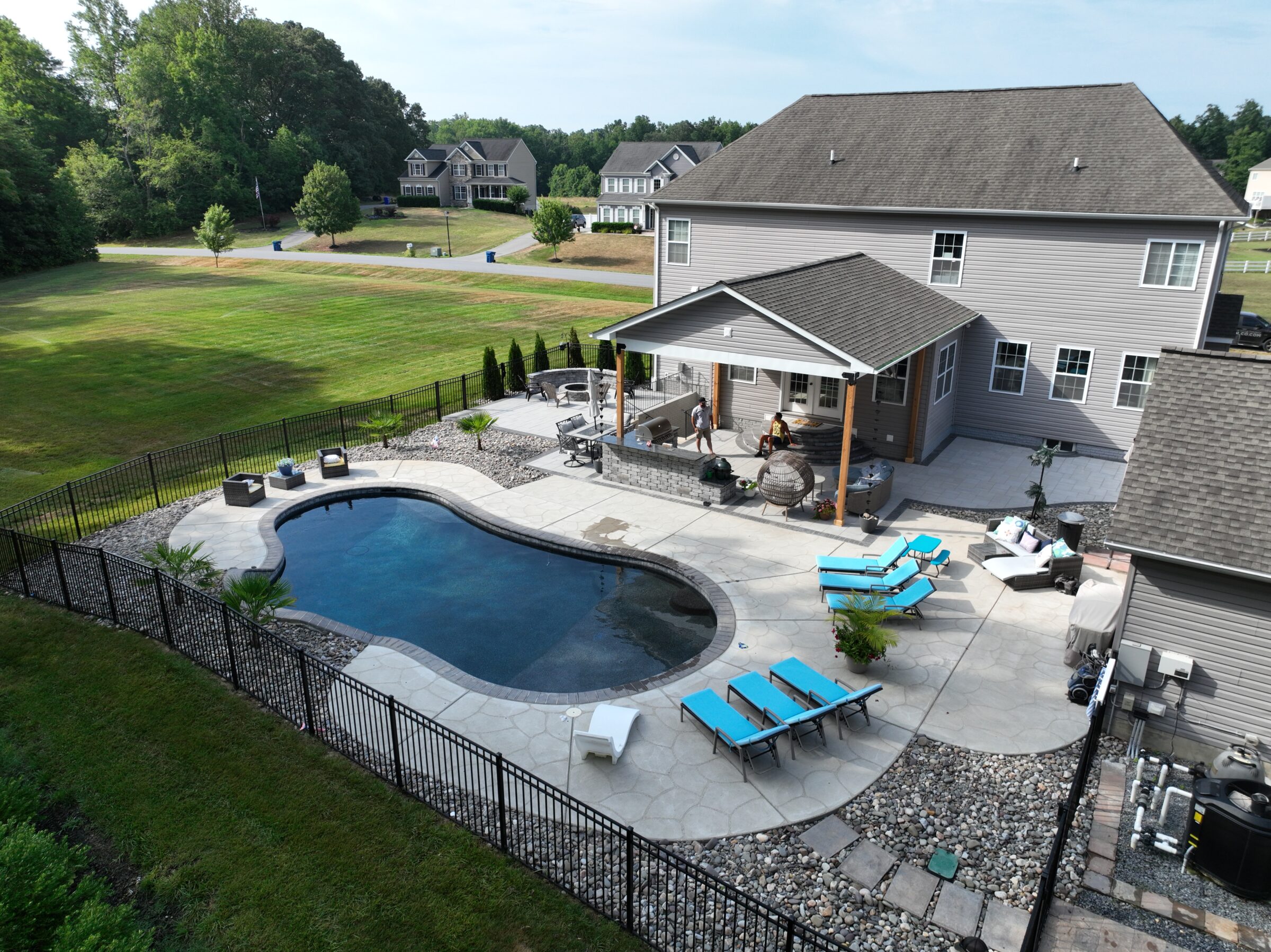 Aerial view of a modern backyard featuring an in-ground pool, lounge chairs with blue cushions, a covered patio area, and landscaped surroundings in a suburban neighborhood.
