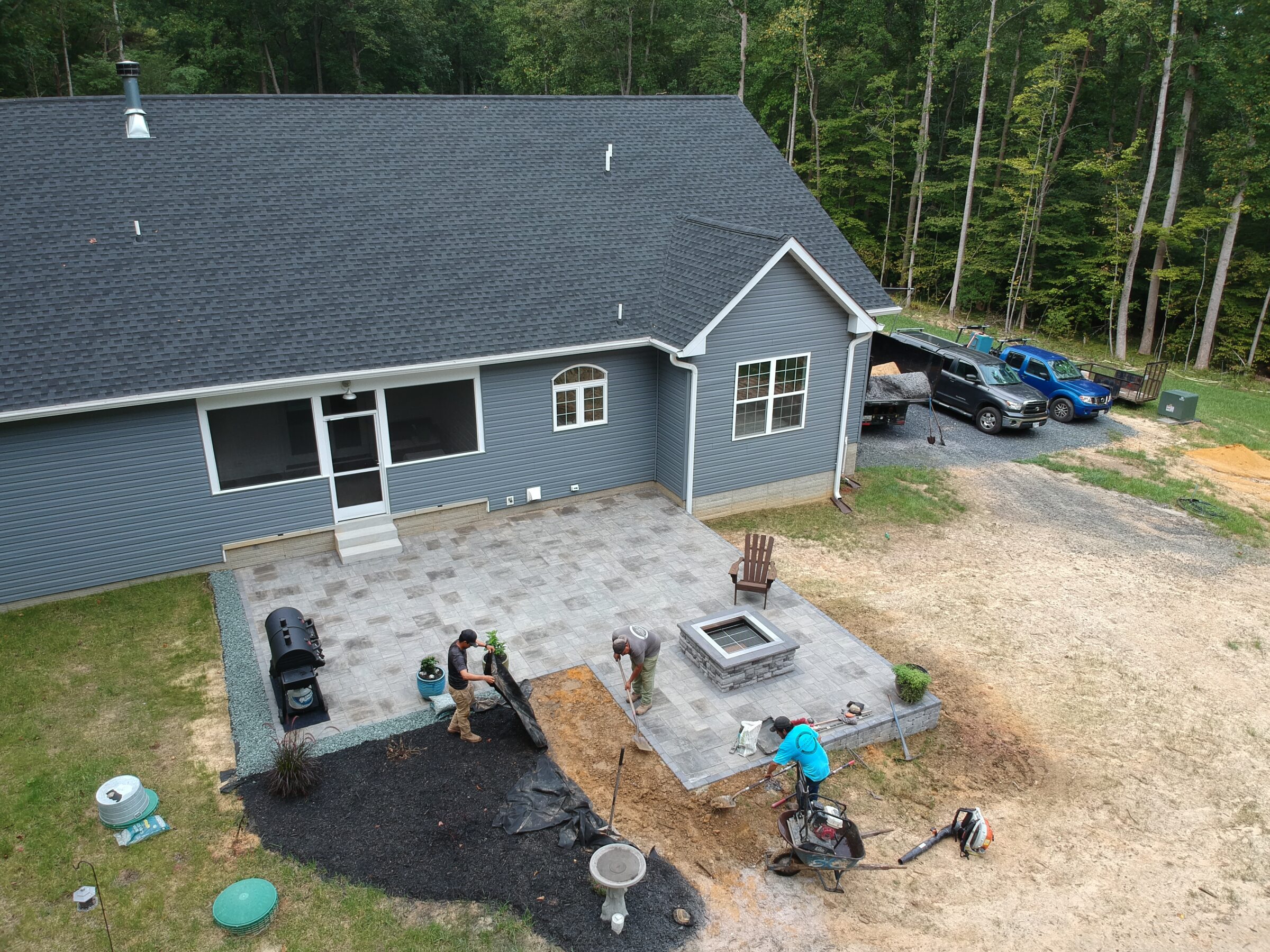 Aerial view of a backyard landscaping project in progress behind a grey house, featuring a newly built stone patio with a fire pit, outdoor seating, and workers installing plants and mulch. Construction vehicles and equipment are visible nearby.