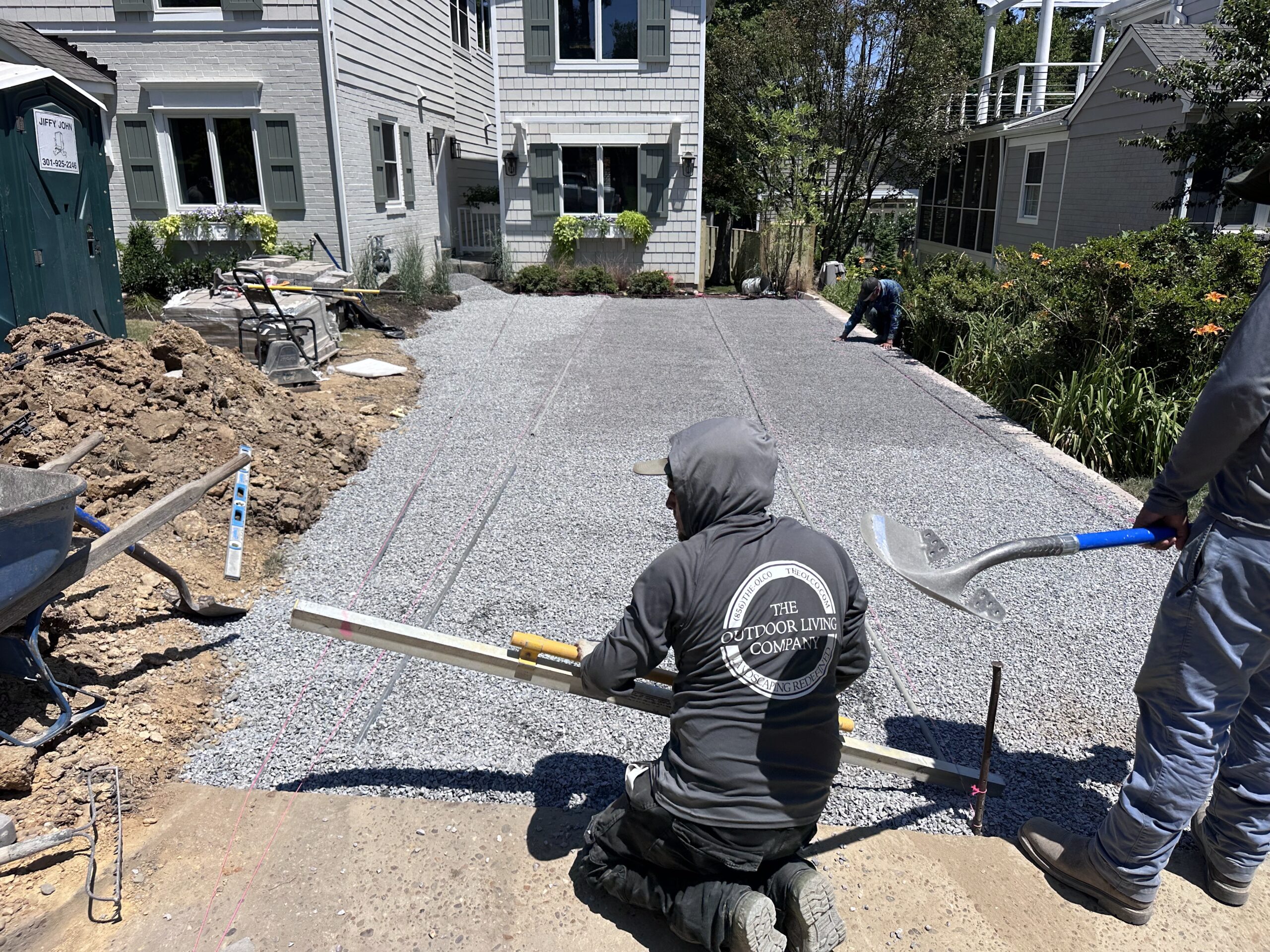 Backyard landscaping project in progress featuring a stone patio with a built-in fire pit, curved retaining wall, and surrounding lawn. Construction materials and tools are visible on-site.