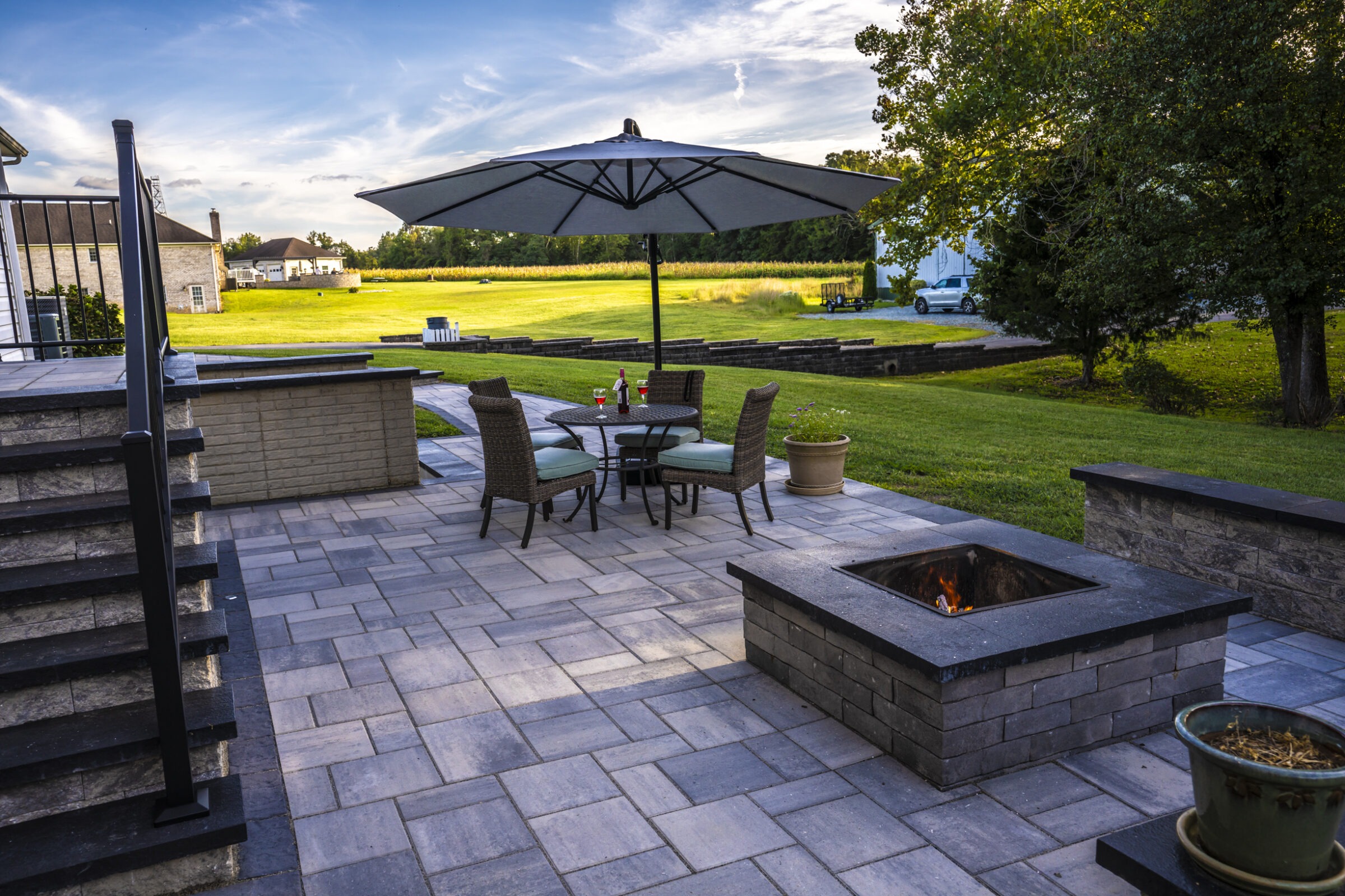 A modern patio with seating, an umbrella, and a fire pit overlooks a spacious lawn, under a clear sky with trees nearby.