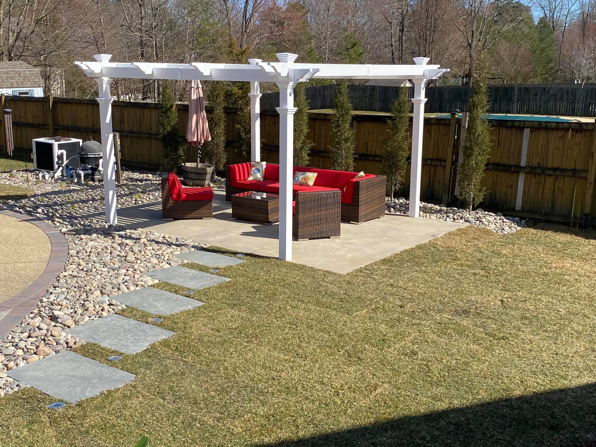 A backyard patio with pergola, red cushions on seating, stone path, and wooden fence, surrounded by grass and trees.