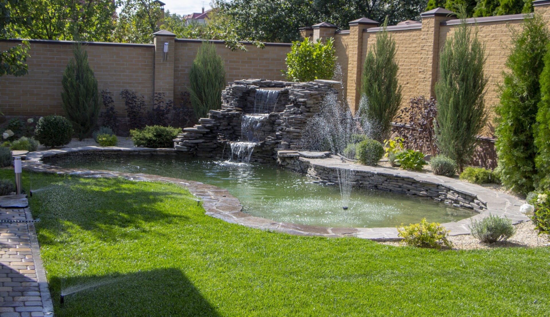 A tranquil garden features a small pond with a stone waterfall and fountains, surrounded by lush greenery and a brick wall.