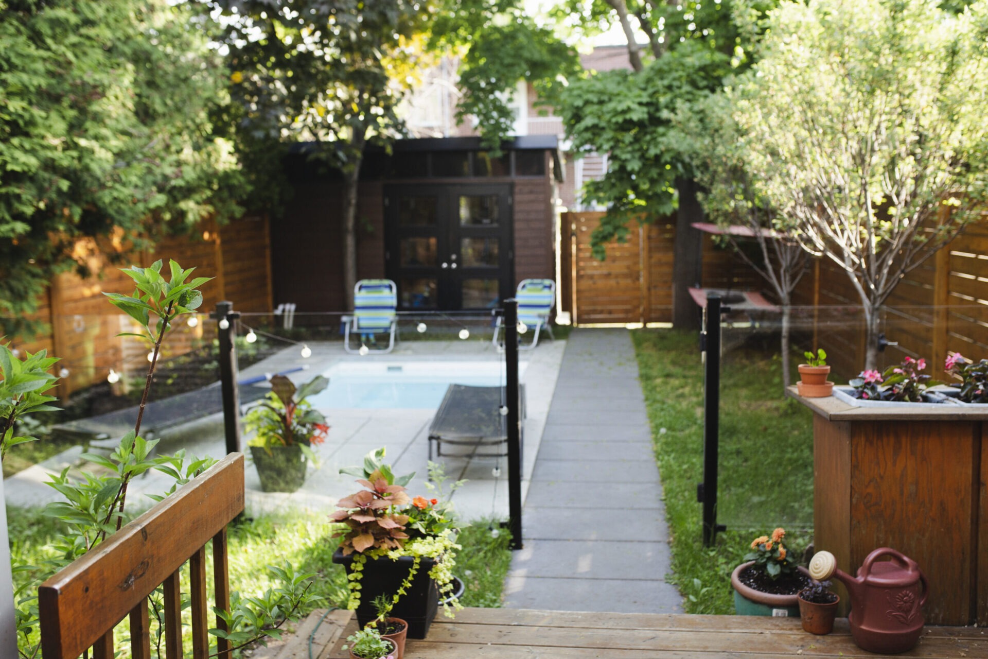 A cozy backyard with a small pool, surrounded by lush greenery, potted plants, and wooden fencing, along with a wooden shed.