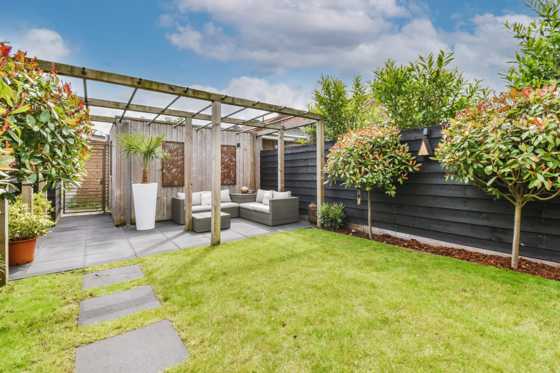 A well-maintained backyard features a pergola, outdoor seating, landscaping with trees, and a lawn under a partly cloudy sky.
