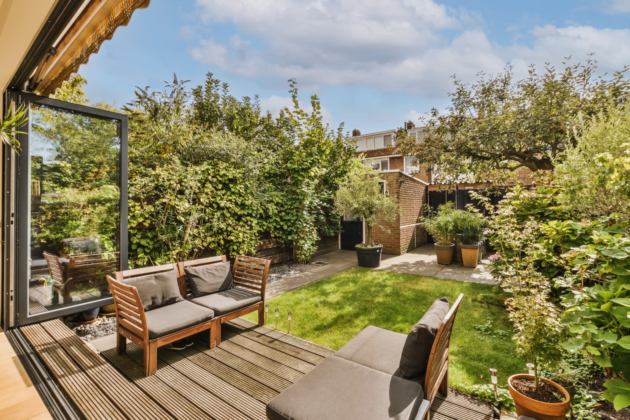 A cozy backyard with wooden patio furniture, lush greenery, potted plants, and an open glass door. Bright and inviting under a blue sky.