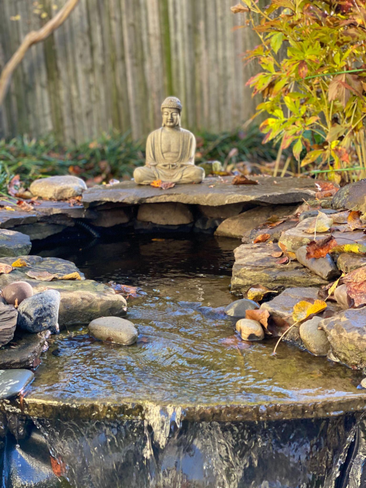Small backyard waterfall with a stone Buddha statue and surrounding rocks and plants.