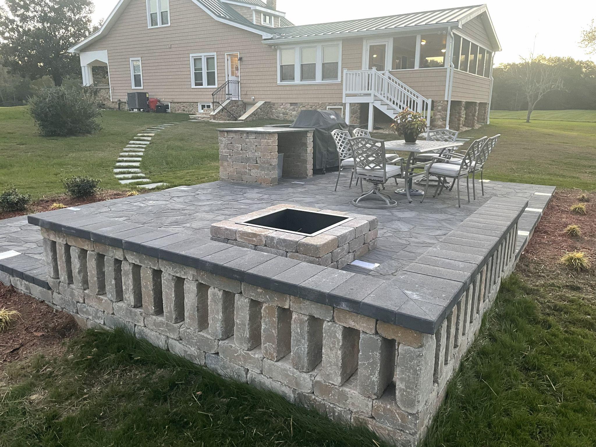 Raised stone patio with a square fire pit and dining set beside a rural home.