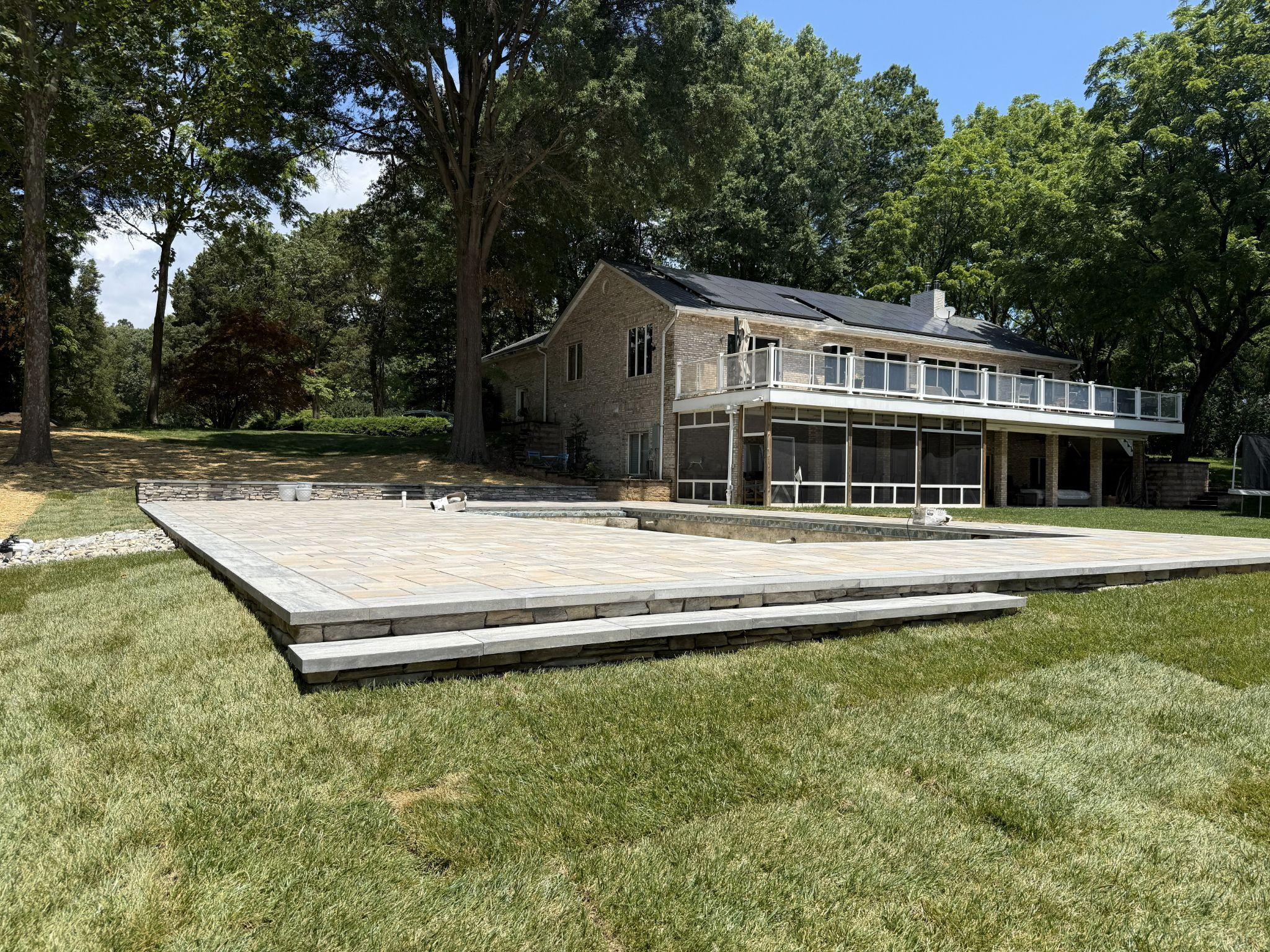 Expansive paver patio under construction behind a large home surrounded by lawn.